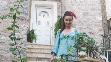 Mavi elbiseli bir kadın balkona iniyor ve sabah serinliğinin tadını çıkarıyor. Gülümsüyor ve çiçeklere bakıyor..