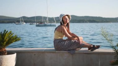 Genç ve çekici bir kadın deniz kenarında bir korkulukta oturmuş manzaranın tadını çıkarıyor. Sarı bir gömlek ve beyaz bir şapka giyiyor..