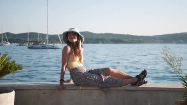 Genç ve çekici bir kadın deniz kenarında bir korkulukta oturmuş manzaranın tadını çıkarıyor. Sarı bir gömlek ve beyaz bir şapka giyiyor..