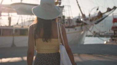 Beyaz şapkalı ve sarı bluzlu bir kadın liman boyunca yelkenliye doğru yürüyor. Beyaz bir çanta taşıyor. Yüksek kalite