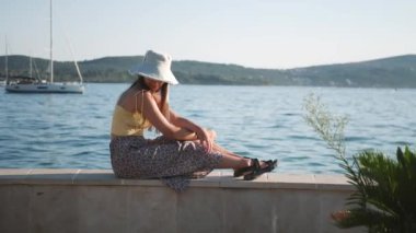 Genç ve çekici bir kadın deniz kenarında bir korkulukta oturmuş manzaranın tadını çıkarıyor. Sarı bir gömlek ve beyaz bir şapka giyiyor..