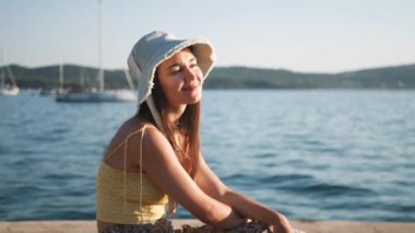 Genç ve çekici bir kadın deniz kenarında bir korkulukta oturmuş manzaranın tadını çıkarıyor. Sarı bir gömlek ve beyaz bir şapka giyiyor..