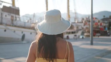 Beyaz şapkalı ve sarı bluzlu bir kadın liman boyunca yelkenliye doğru yürüyor. Beyaz bir çanta taşıyor. Yüksek kalite