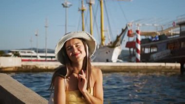 Bir kadın su kenarındaki bir çıkıntıda oturuyor, üzerinde sarı bir bluz ve beyaz bir şapka var. Hava öpücüğü yolluyor. Yüksek kalite