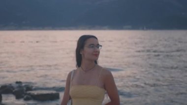 Genç ve çekici bir kadın sahilde duruyor ve körfez ve dağların güzel manzarasının tadını çıkarıyor. Yüksek kalite