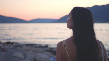 Yüzünde bir gülümsemeyle sahilde duran bir kadın. O, sarı bir elbise ve bir kolye giyiyor. Arka planda bir günbatımı, bir okyanus ve muhteşem bir dağ manzarası var. Yüksek