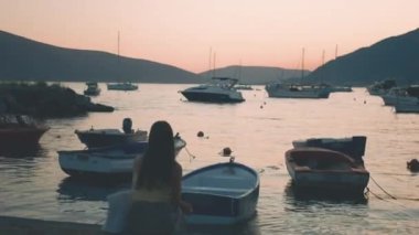 Bir kadın sahilde teknenin yanında oturur ve günbatımının tadını çıkarır. Su sakin ve arka planda birkaç tekne var. Sahne huzurlu ve huzurlu. Yüksek kalite