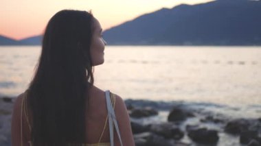 Yüzünde bir gülümsemeyle sahilde duran bir kadın. O, sarı bir elbise ve bir kolye giyiyor. Arka planda bir günbatımı, bir okyanus ve muhteşem bir dağ manzarası var. Yüksek