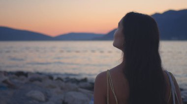 Genç ve çekici bir kadın sahilde duruyor ve körfez ve dağların güzel manzarasının tadını çıkarıyor. Yüksek kalite