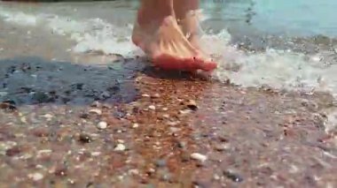 Sahilde kayalık bir sahilde genç bir kızın bacakları. Bir kadın deniz kıyısında yalınayak yürür ve dalgalar ayaklarını yıkar. Yüksek kalite