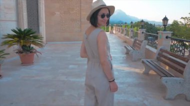  Genç çekici bir kız cadde boyunca yürür. Beyaz şapkalı genç bir kız cadde boyunca yürüyüp güzel bir manzaranın tadını çıkarıyor..