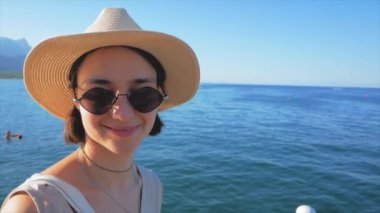 Genç ve çekici bir kızın portresi. Beyaz şapkalı ve güneş gözlüklü çekici bir kız kameraya bakıyor ve gülümsüyor. Genç milenyum kızı harika bir tatilden zevk alıyor. Yüksek kalite 4k görüntü