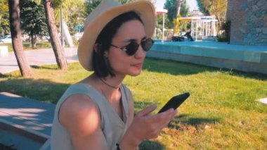 Sosyal medyada akıllı telefonuyla gezinen genç ve çekici bir kızın portresi. Beyaz şapkalı ve güneş gözlüklü çekici bir kız kameraya bakıp gülümsüyor. Genç bir milenyum