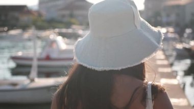 Sarı elbiseli ve mavi şapkalı bir kadın birkaç teknesi olan bir rıhtımın önünde duruyor. Genç, çekici, tasasız bir kız rıhtımda yürüyor..