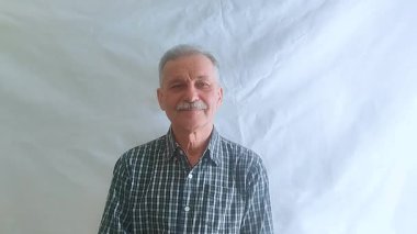 Bıyıklı ve kareli gömlekli yaşlı bir adam hayretle kameraya bakar. Beyaz arkaplanda bir portre.