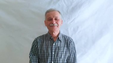 Bıyıklı ve kareli gömlekli yaşlı bir adam kameraya bakarken gülüyor. Beyaz arkaplanda bir portre.