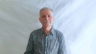 Bıyıklı ve kareli gömlekli yaşlı bir adam kameraya bakar. Beyaz arkaplanda bir portre.