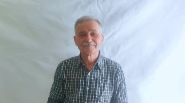 Hevesli bir ruh hali içinde yaşlı bir adam elini kibarca sallıyor ve kameraya bakıyor. Beyaz arkaplanda bir portre.