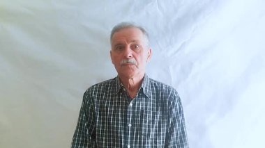 Bıyıklı ve kareli gömlekli yaşlı bir adam hayretle kameraya bakar. Beyaz arkaplanda bir portre.