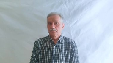 Bıyıklı ve kareli gömlekli yaşlı bir adam hayretle kameraya bakar. Beyaz arkaplanda bir portre.