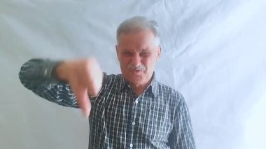 Yaşlı bir adam üzgün bir ruh hali içinde baş parmağını eğiyor, başını negatif bir şekilde sallıyor ve kameraya bakıyor. Beyaz arkaplanda bir portre.