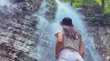 Çekici bir genç kız turist muhteşem bir dağ şelalesinin önünde duruyor ve onun büyüklüğünün tadını çıkarıyor. Çekici bir gezgin kız muhteşem bir şelaleye bakıyor. Yüksek kalite 4k görüntü