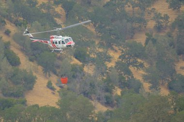 Tepelerde elinde su kovasıyla uçan bir Cal Fire helikopteri. Berryessa Gölü, Kaliforniya, ABD