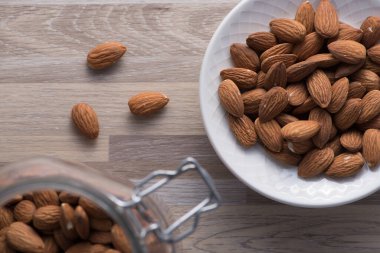 Beyaz kase dolusu çiğ badem, rustik ahşap yüzey kopça kavanozu dolu.