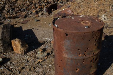 Paslı metal fıçının kirli zemine yerleştirilmiş delikler ve güneş ışığında kayalar ve çöpler. 