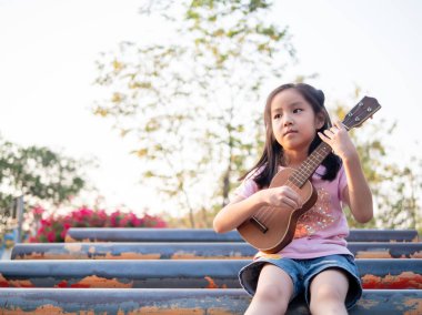 Küçük Asyalı kız ukulele çalıyor, çelik boruyla bahçede.