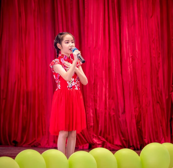 The Asian kid girl sing a song on stage at her school activity day, dress in Qipao style, red curtain background