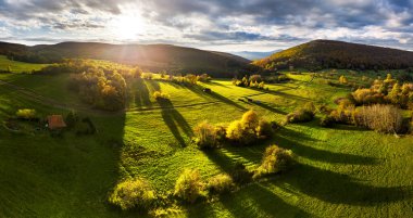 Wonderful autumn colored forest, setting sun with dramatic sky, valley with green grass and trees. Sunlight nature landscape clipart