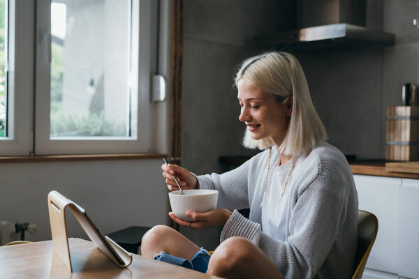 young adult caucasian woman having breakfast and using digital tablet in her kitchen