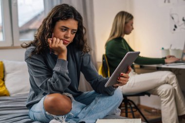 Dijital tablet kullanan bayan üniversite öğrencisi yataktaki yurt odasında oturuyor.