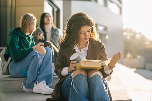 キャンパスの屋外で本を読んでいる若い大人の女性の肖像画です 背景がぼやけている友人は — ストック写真