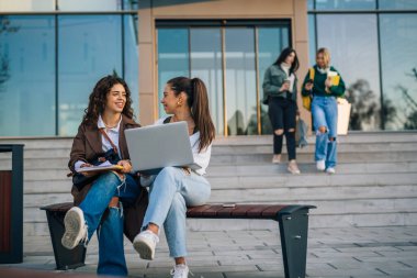 İki üniversite öğrencisi fakültenin önündeki bankta oturuyor.