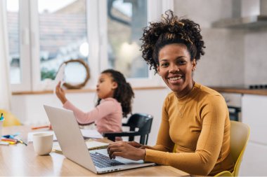 Kızı arka planda oynarken melez anne evde dizüstü bilgisayarda çalışıyor.