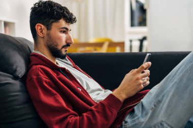 Kanepede oturan ve telefonunu kullanan adamın yan görüntüsü.