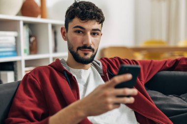 Genç yetişkin bir adam evde oturup kameraya bakıyor.