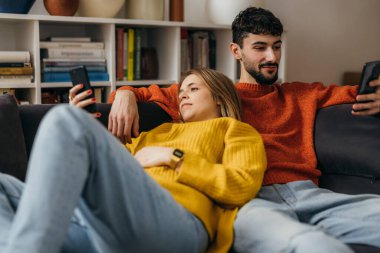 Genç çift kanepede yatar ve telefona bakar.