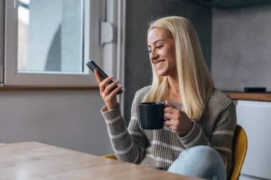 Beautiful woman looks at her phone and enjoys coffee in the kitchen