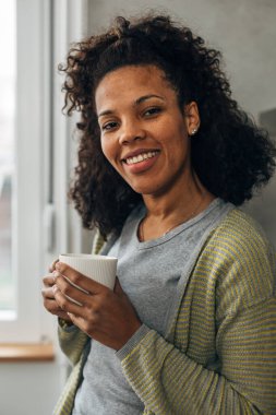 Melez bir kadın elinde fincanla kameraya gülümsüyor.