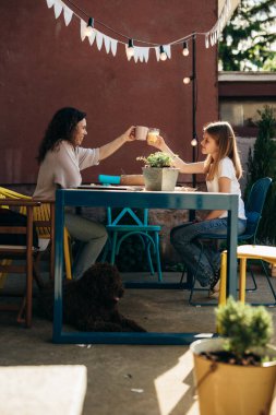 W kadın ve kızı arka bahçede kahvaltı ederken bardak tokuştururlar.