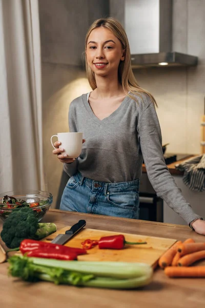 stock image A young blond woman is standing in he kitchen with a cup of coffee and cooks a vegan meal