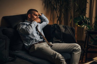 İşten eve bir işadamı geldi. Kanepeye oturur ve sıcak içeceği tek başına içer.