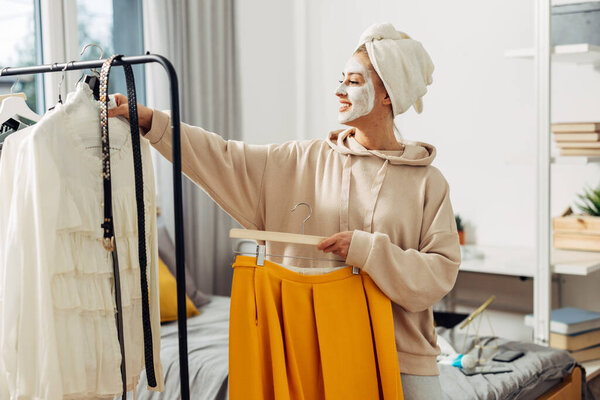 Счастливая женщина в маске и полотенце тюрбан выбирает одежду, чтобы носить