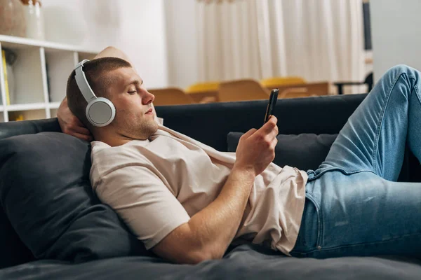 一个白人拿着智能手机躺在沙发上的侧视图 — 图库照片