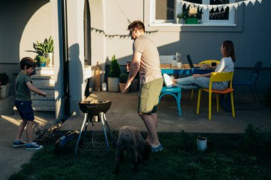 Aile güneşli bir yaz gününde bahçededir. Baba ve oğul ızgaraya odun koyup barbekü için ateş yaktılar.