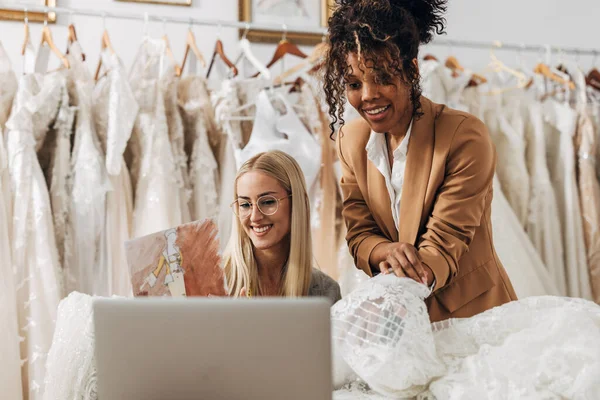 Two women work in a bridal tailoring salon and talking on a video call with a client to choose material for a wedding dress.