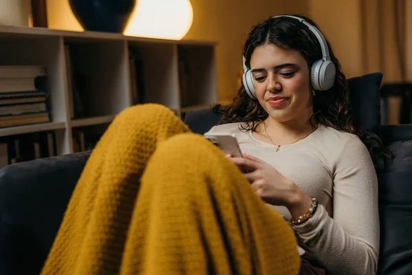 stock image A cute young woman is sitting alone on the sofa with a blanket and using her phone.